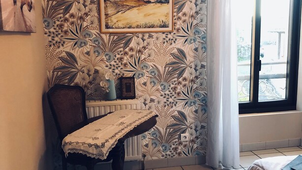 A room with a patterned wallpaper, a framed landscape painting, a table with a lace tablecloth, a chair, a radiator, and a window with curtains. - ROOM for 2 adults 1 h30 from TOULOUSE - Pets under -3 kg accepted. (SAINT-PROJET)