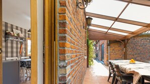 Een overdekt terras met een bakstenen muur, een eettafel met stoelen, en een keukenhoek met een geruit wand en hangende objecten.