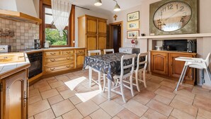 Een keuken met houten kastjes, een eettafel met stoelen, een grote wandklok en een raam met gordijnen.