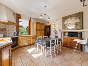 Une cuisine avec des meubles de rangement en bois, une table à manger avec des chaises, une grande horloge murale et une fenêtre avec des rideaux.