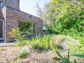 A modern brick building with a large window, surrounded by a garden with various plants and a pathway. - Comfortable house in the heart of the village (Le Bizet)