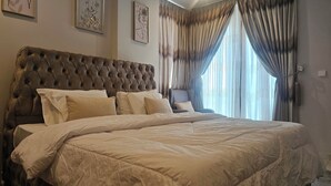 Una cama grande con cabecera capitonada, varios cojines y una colcha de color claro. Hay una silla y una ventana con cortinas.