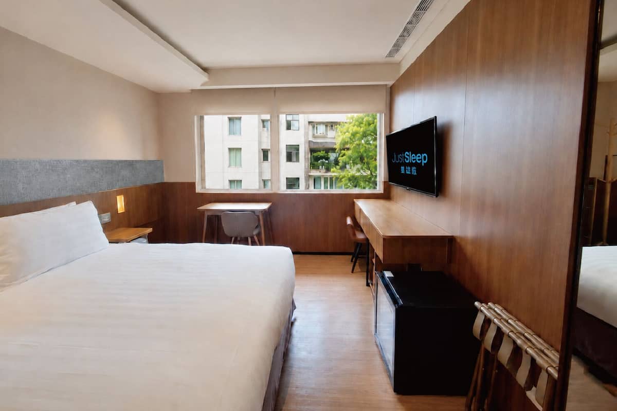 A hotel room with a bed, a desk, a chair, a TV, and a window with a view of buildings and greenery.