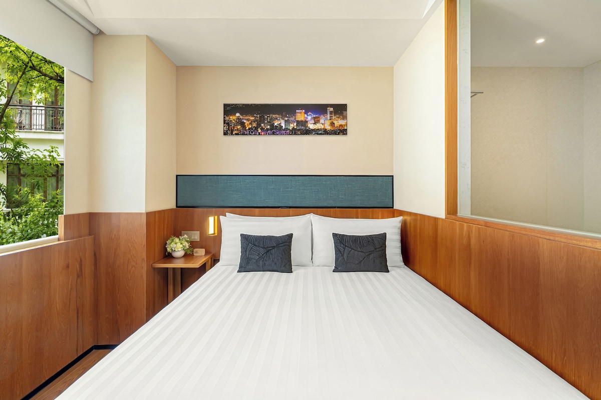 A hotel room with a bed, a nightstand, a painting, and a view of greenery.