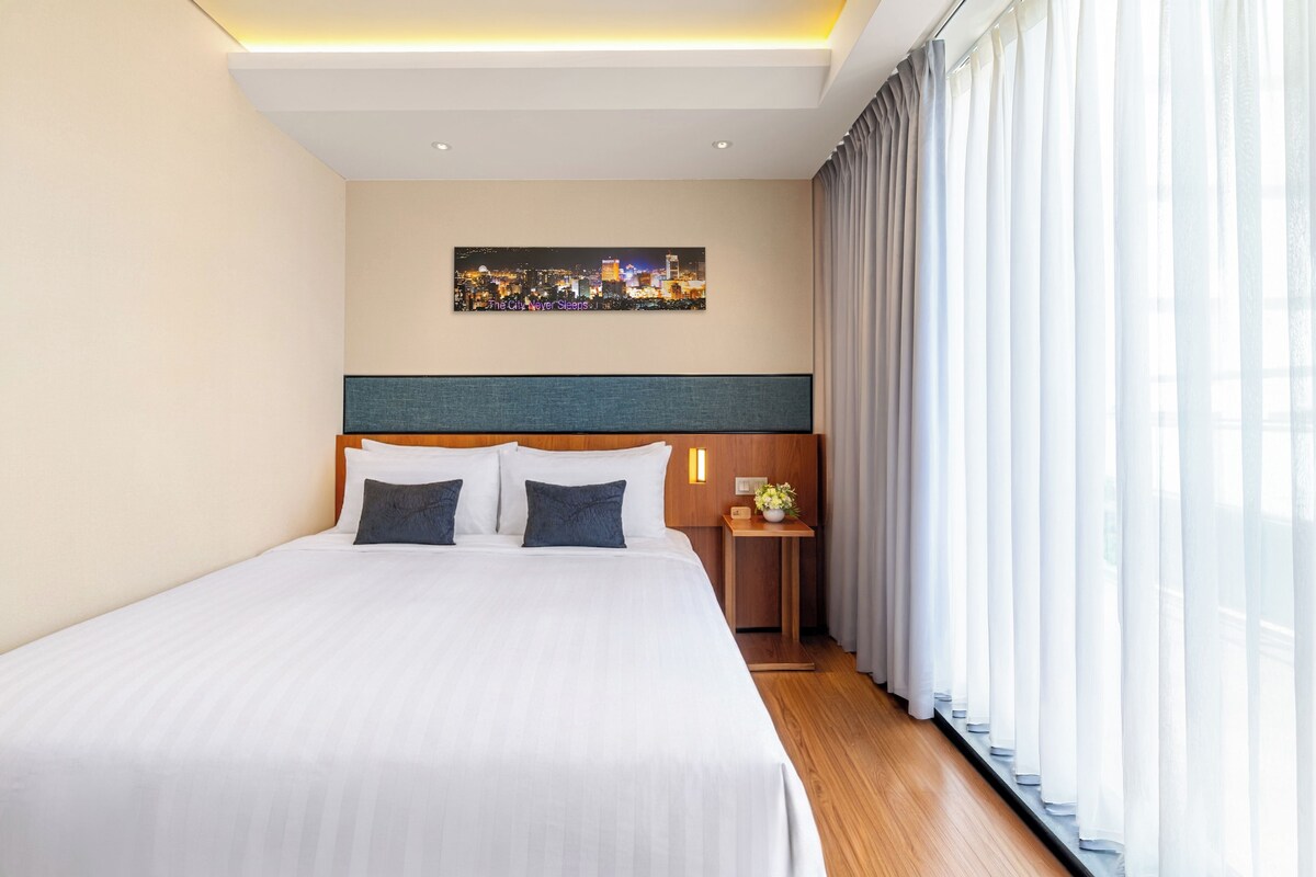 A hotel room with a large bed, a nightstand with a lamp, a painting on the wall, and sheer curtains.