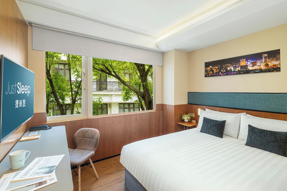 Une chambre d’hôtel comprenant un lit, un bureau avec une chaise, une télévision et une fenêtre donnant sur des arbres et des immeubles.