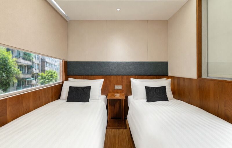 Quarto de hotel com duas camas, cabeceira de madeira, uma mesa pequena e janela com vista para prédios.