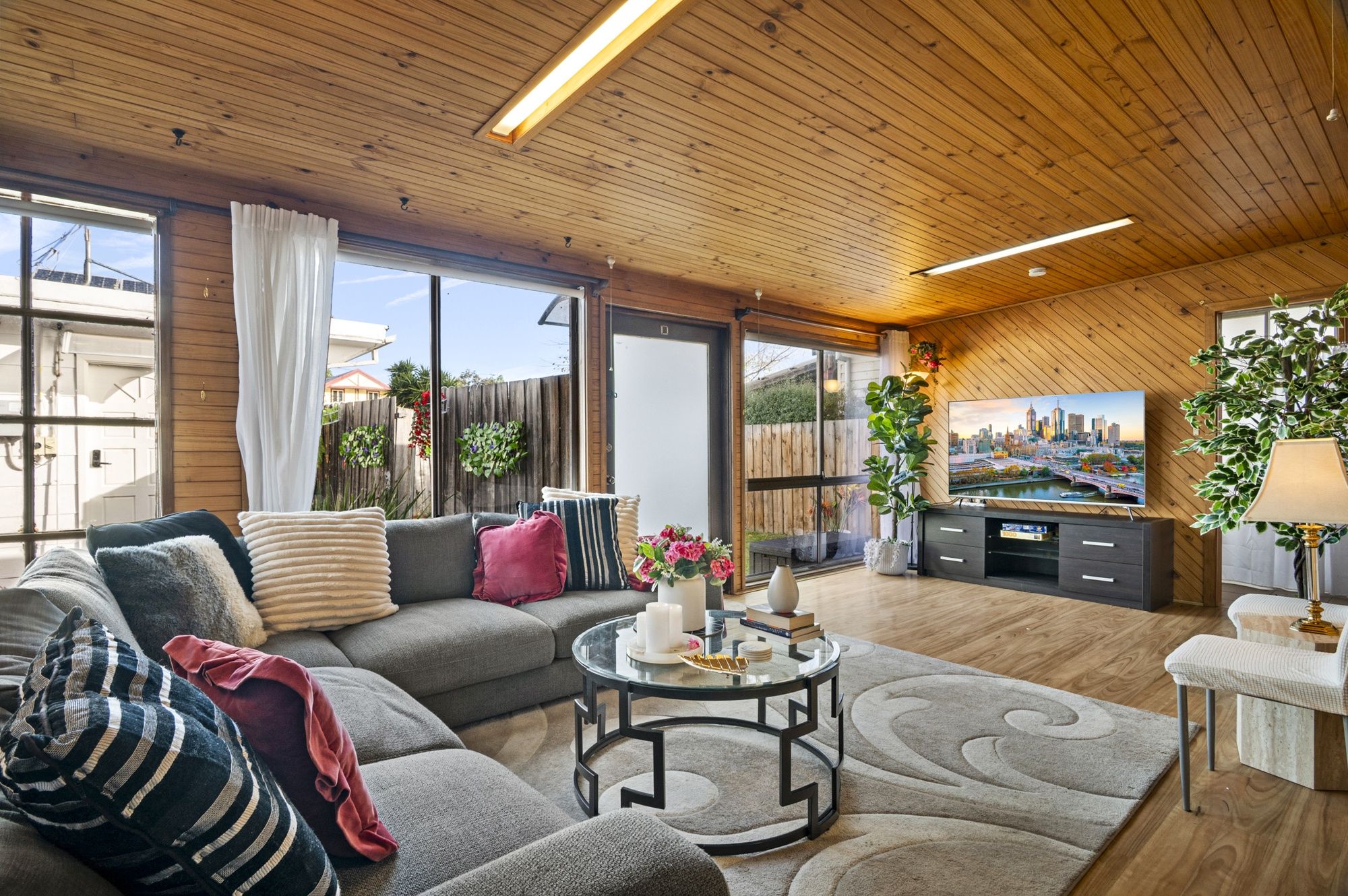 A modern living room with a sectional sofa, a glass coffee table, and a flat-screen TV mounted on a wooden wall.