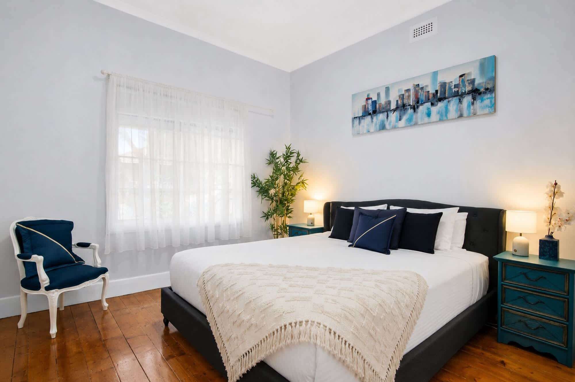 A bedroom with a bed, a chair, a window with sheer curtains, a cityscape painting, and a plant.