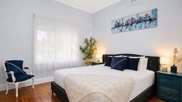 A bedroom with a bed, a chair, a window with sheer curtains, a cityscape painting, and a plant.