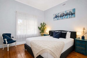 A bedroom with a bed, a chair, a window with sheer curtains, a cityscape painting, and a plant. - Spacious 4-Bed Retreat in Vibrant Footscray (Footscray)