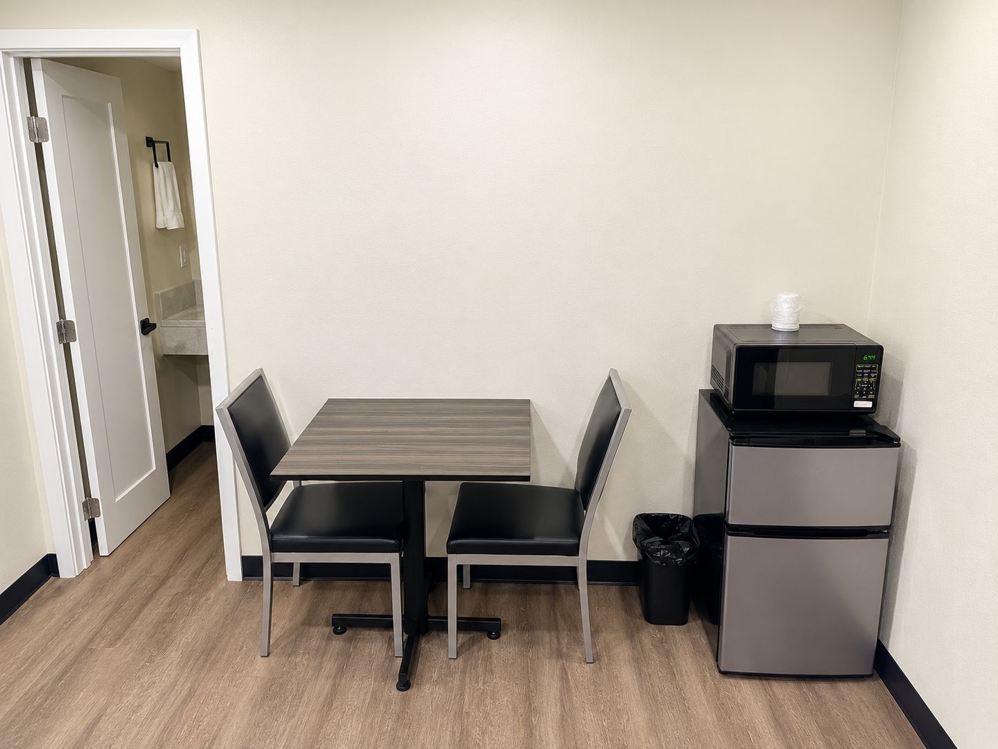 Una kitchenette pequeña con microondas y refrigerador, una mesa con dos sillas, y una puerta que da a un baño.