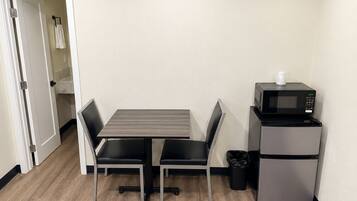 Una kitchenette pequeña con microondas y refrigerador, una mesa con dos sillas, y una puerta que da a un baño.