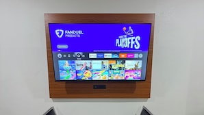 Un televisor instalado en la pared mostrando una interfaz de servicio de streaming con un banner que dice 'PANDUEL PREDICTS PLAYOFFS'.