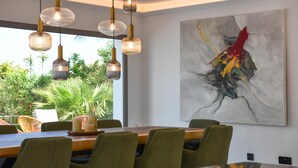 Ein Essbereich mit einem großen Tisch, grünen Polsterstühlen, Pendelleuchten und einem großen abstrakten Gemälde an der Wand.