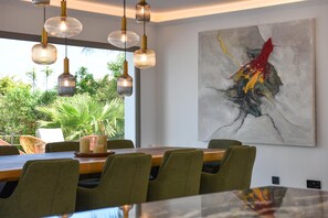 A dining area with a large table, green upholstered chairs, pendant lighting, and a large abstract painting on the wall. - Villa Amantos 8 pax - Denia, Costa Blanca (Denia)