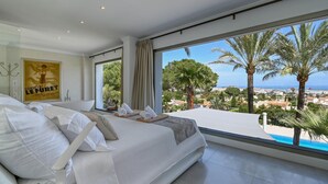 Ein Schlafzimmer mit einem großen Bett, Blick auf Palmen und das Meer, und einem Poster an der Wand.