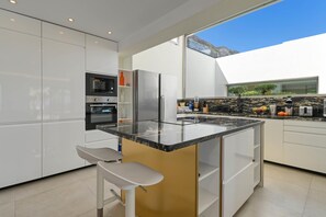 A modern kitchen with white cabinets, a central island with seating, a built-in oven, and a large window offering a view of the outdoors. - Villa Amantos 8 pax - Denia, Costa Blanca (Denia)