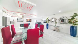 A modern dining area with a glass table and red chairs, a kitchen in the background, and decorative wall art.