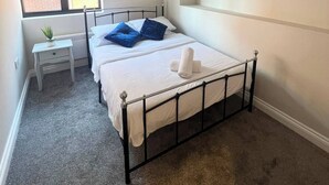 Et soveværelse med en enkelt seng, et lille natbord, en potteplante og et vindue med udsigt til en murstensbygning.