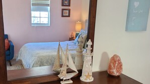 Un dormitorio con cómoda de madera, espejo que refleja la habitación, cama con colcha blanca, mesita de noche con lámpara, y pared con un cuadro enmarcado y una cita motivacional.