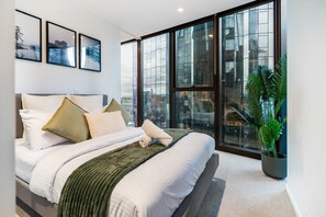 Ein modernes Schlafzimmer mit einem großen Bett, Blick über die Stadt durch raumhohe Fenster und einer Topfpflanze.