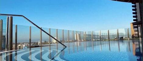 A clear blue sky, a swimming pool with a glass barrier, and a cityscape in the background.