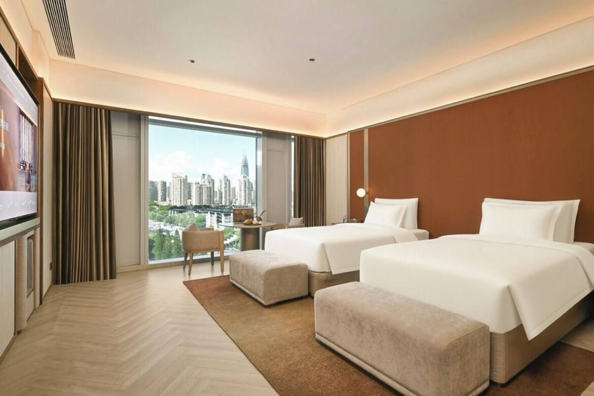 Quarto de hotel com duas camas, televisão, escrivaninha e uma grande janela com vista para a cidade.