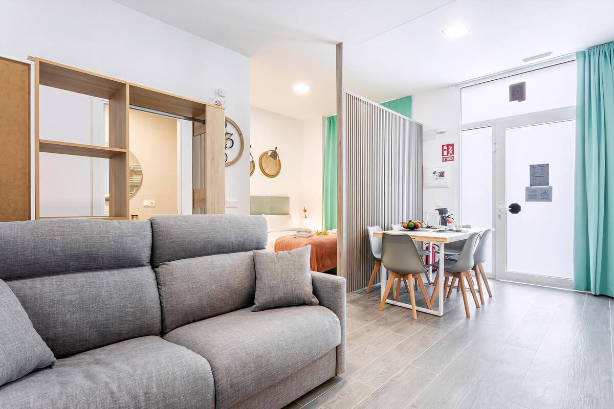 A modern living room with a grey sofa, a dining area with a table and chairs, and a kitchen in the background.