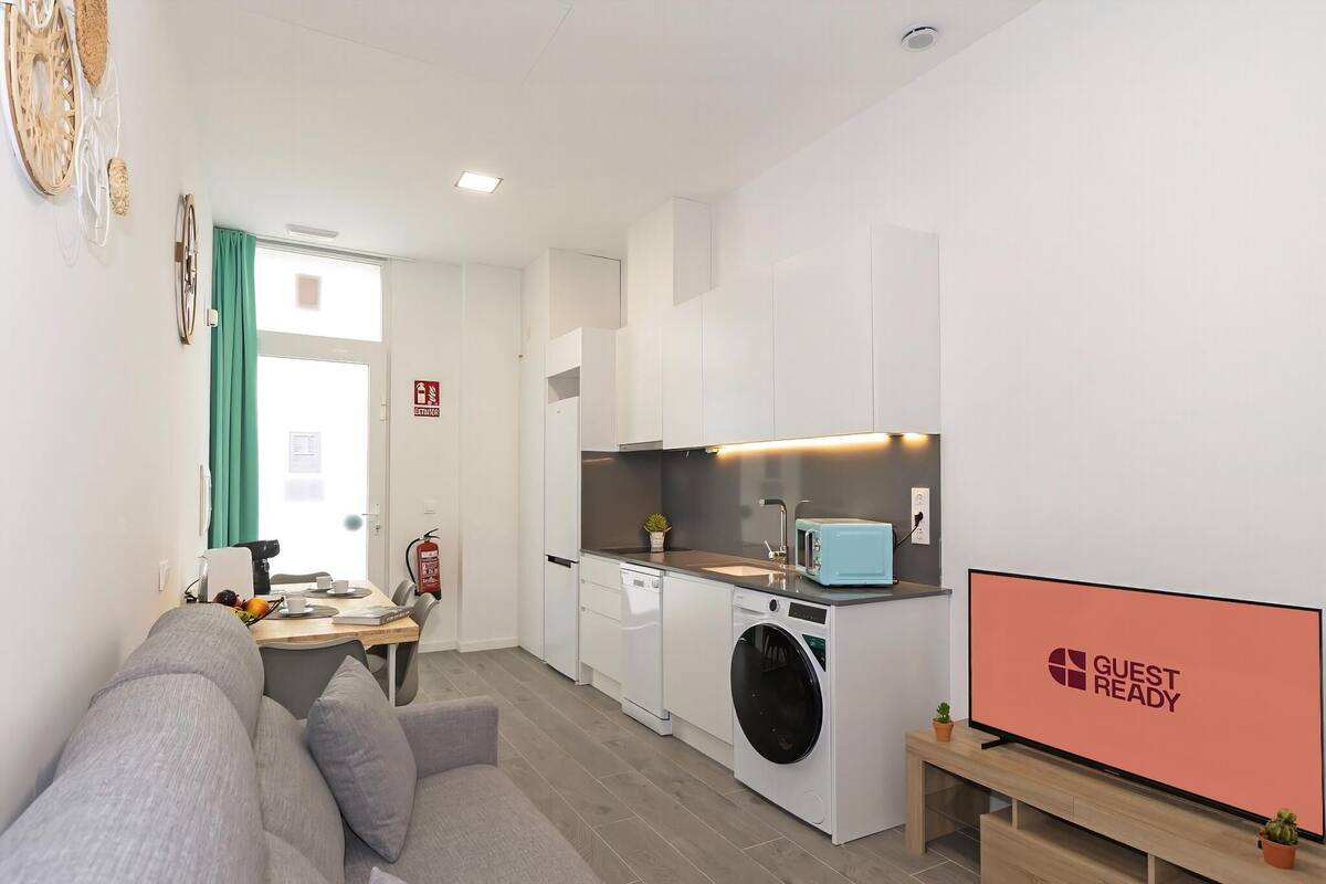 A modern kitchen with a washing machine, a dining area with a table and chairs, and a flat-screen TV displaying 'GUEST READY'.