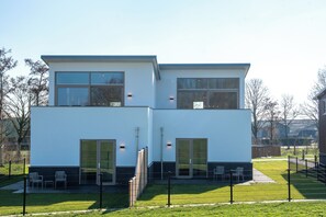 Un bâtiment moderne à deux étages, doté de grandes fenêtres, d’un toit plat et d’un espace extérieur clôturé aménagé avec des chaises.
