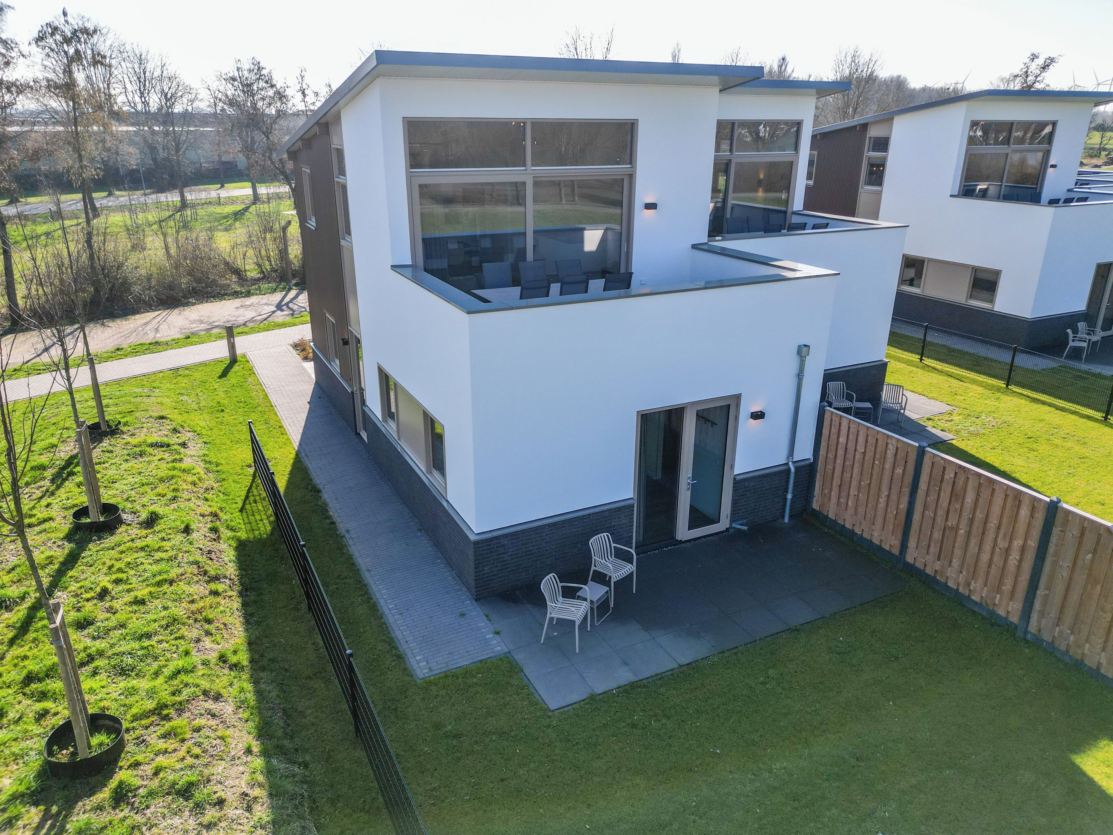 Una casa moderna de dos pisos con techo plano, ventanas grandes y un patio con mobiliario exterior.