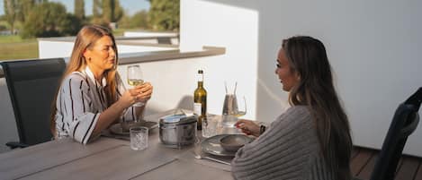 Twee personen zitten aan een buitentafel met een fles wijn, glazen en een pot.