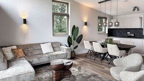 Un salon moderne comprenant un canapé d’angle, une table à manger et des chaises, une grande fenêtre et un coin cuisine avec des luminaires suspendus.