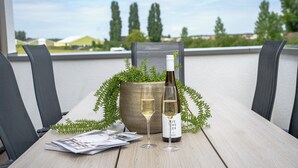 A table with a bottle of wine, two glasses, magazines, and a potted plant on a balcony with a view of trees and buildings. - Experience the ultimate holiday feeling in a stylish waterfront holiday home, with the beach within walking distance.<br>Located close to the Hoge Veluwe National Park and the beautiful Posbank – ideal for nature lovers, hikers, and cyclists. (Giesbeek)