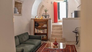 Una sala de estar acogedora con un sofá verde, una estantería de madera, una mesita de centro con superficie de vidrio y una escalera que sube a un nivel superior.