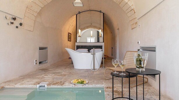 Una piscina interior con una zona de estar moderna, una cocina con electrodomésticos blancos y una mesa de comedor con vasos y una botella.