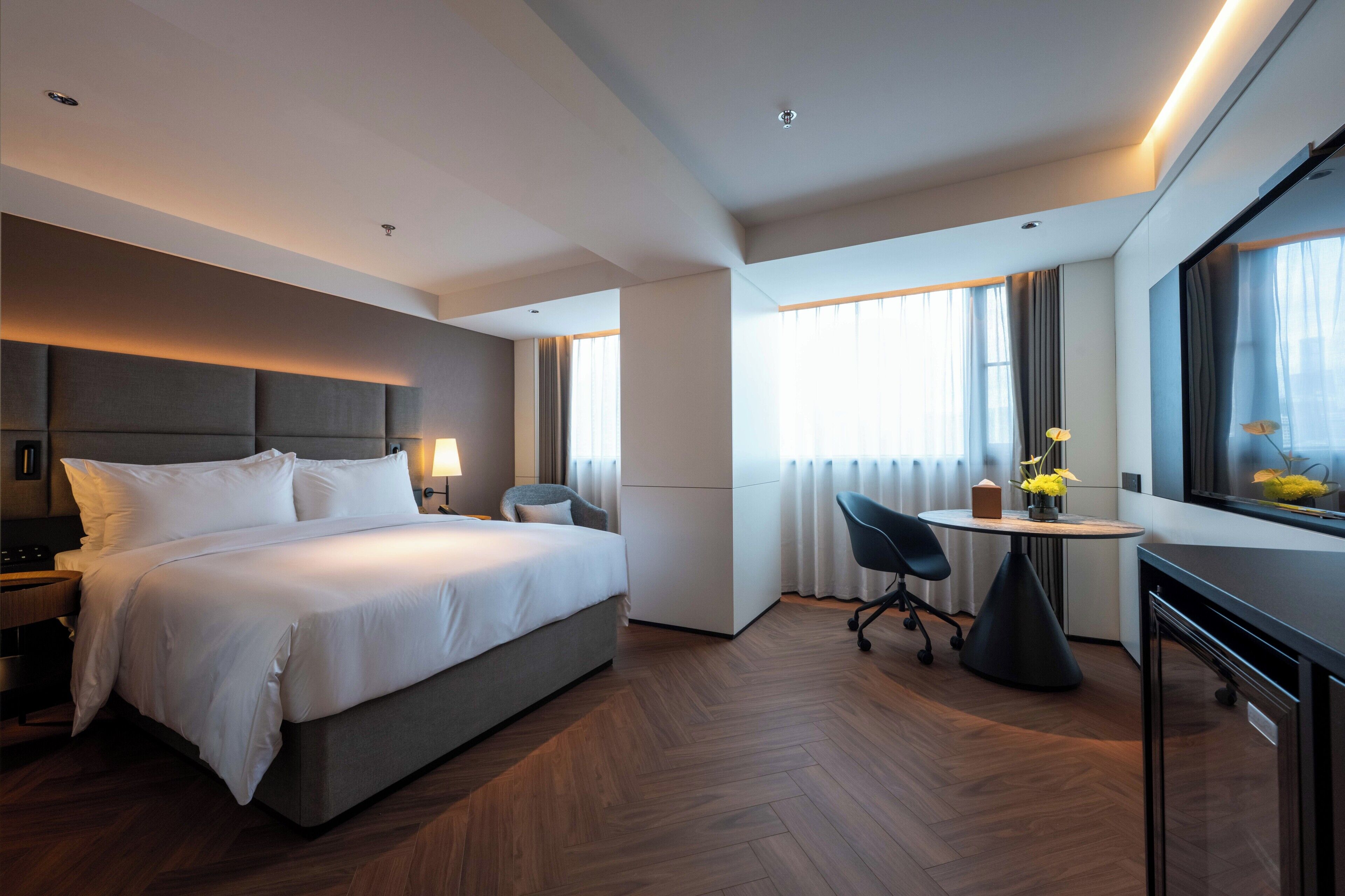 Habitación de hotel moderna con una cama grande, un escritorio, una silla, una pequeña mesa con flores y un ventanal con cortinas.