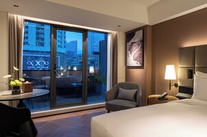 A hotel room with a large window, a bed, a chair, a small table with a lamp, and a wall-mounted phone.