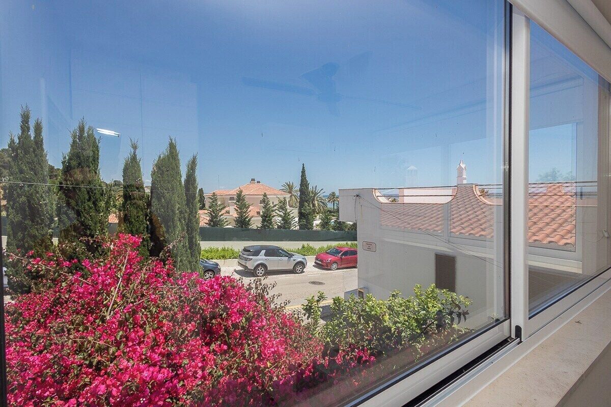 Vista chiara di un parcheggio con diverse auto, un edificio con tetto in tegole rosse e una fila di alberi alti.