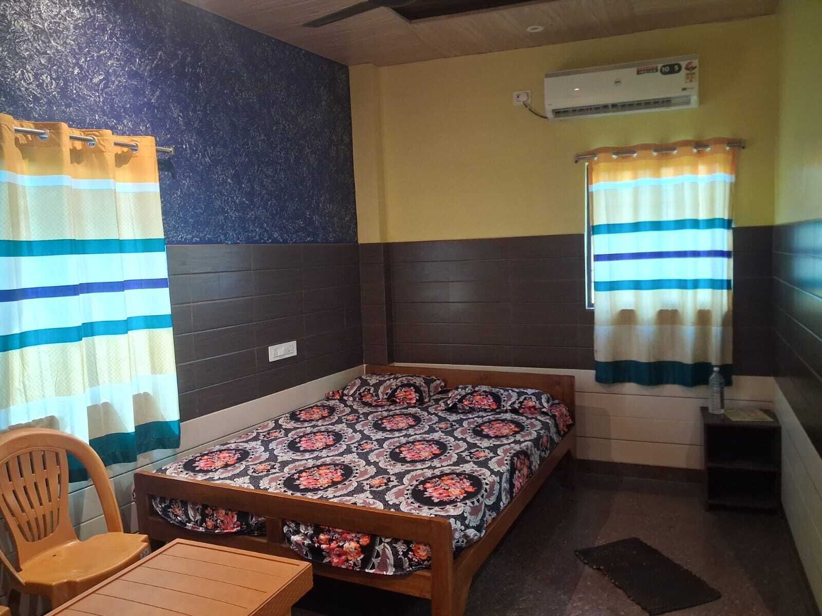 Un dormitorio con cama, una silla, una mesita, un equipo de aire acondicionado y cortinas.