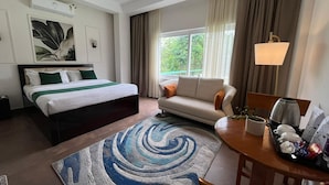 Et hotelværelse med seng, sofa, spisebord med bakke, lampe og udsigt til grøn vegetation udenfor.