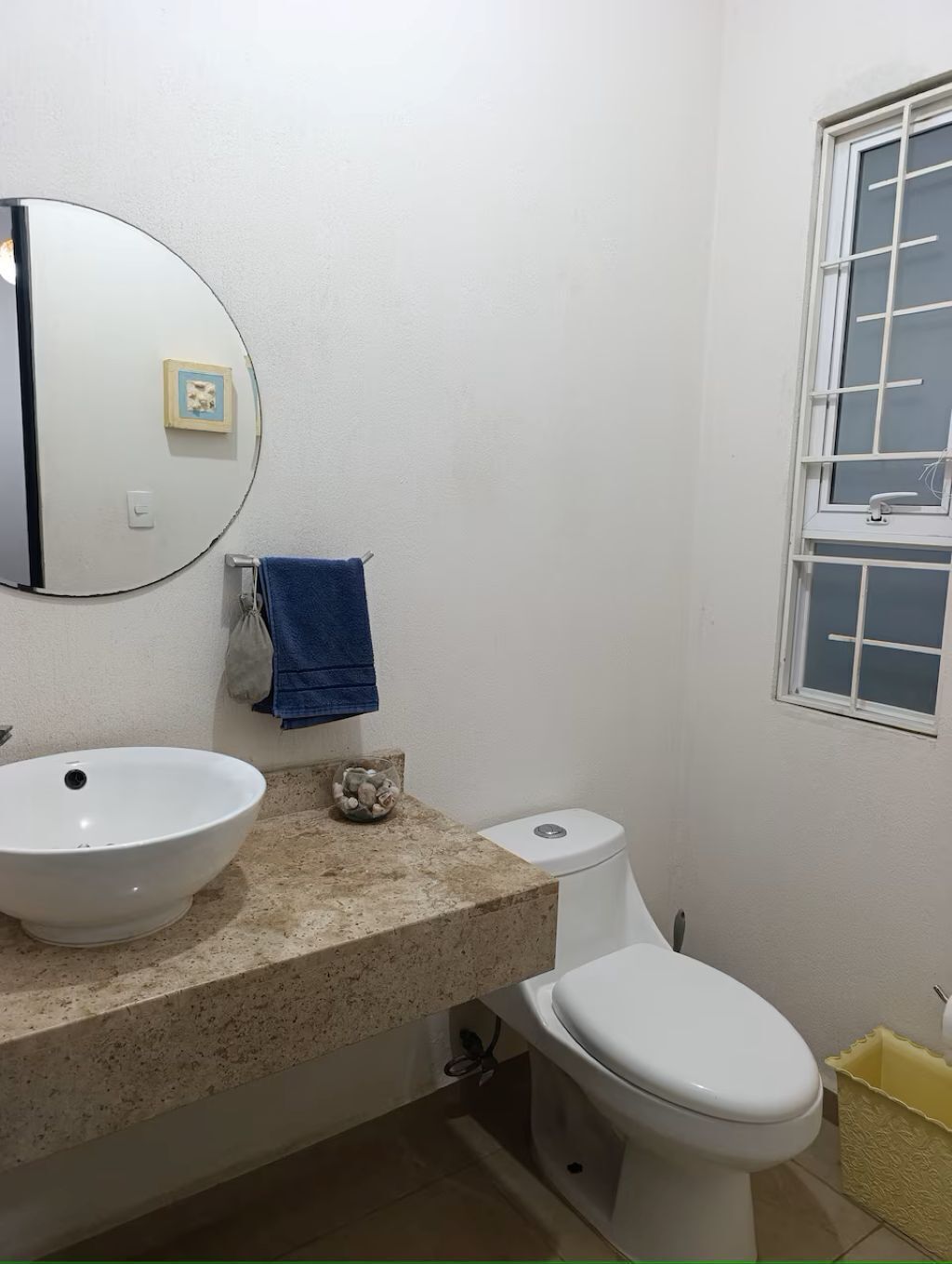 A bathroom with a round mirror, a sink, a toilet, and a window.