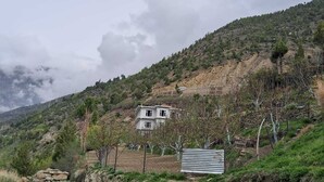 Une maison de campagne, accessible par un chemin de terre, entourée d’arbres et de collines.