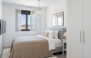 Ein Schlafzimmer mit Bett, Nachttisch, Lampe, Kleiderschrank, einem Fenster mit Vorhängen und einem gerahmten Bild an der Wand.