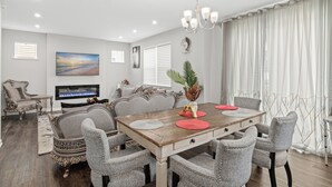 A dining area with a wooden table, grey upholstered chairs, a chandelier, and a flat-screen TV mounted on the wall. - A comfortable 3-bedroom home offers 5 beds and can accommodate up to 8 guests. (Rogers)