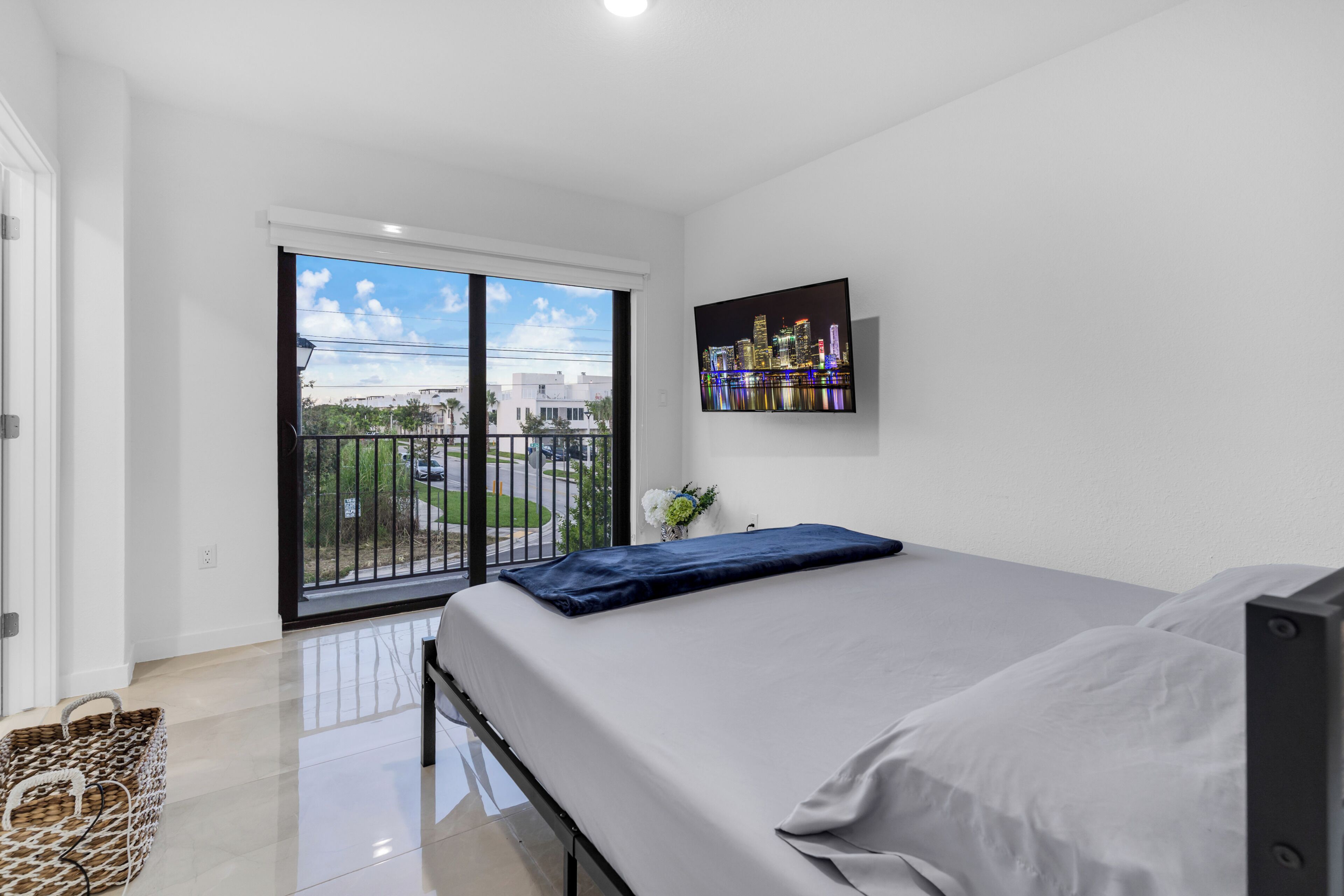 A bedroom with a bed, a cityscape painting, and a balcony with a view of buildings and greenery.