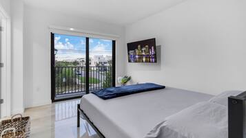 A bedroom with a bed, a cityscape painting, and a balcony with a view of buildings and greenery.
