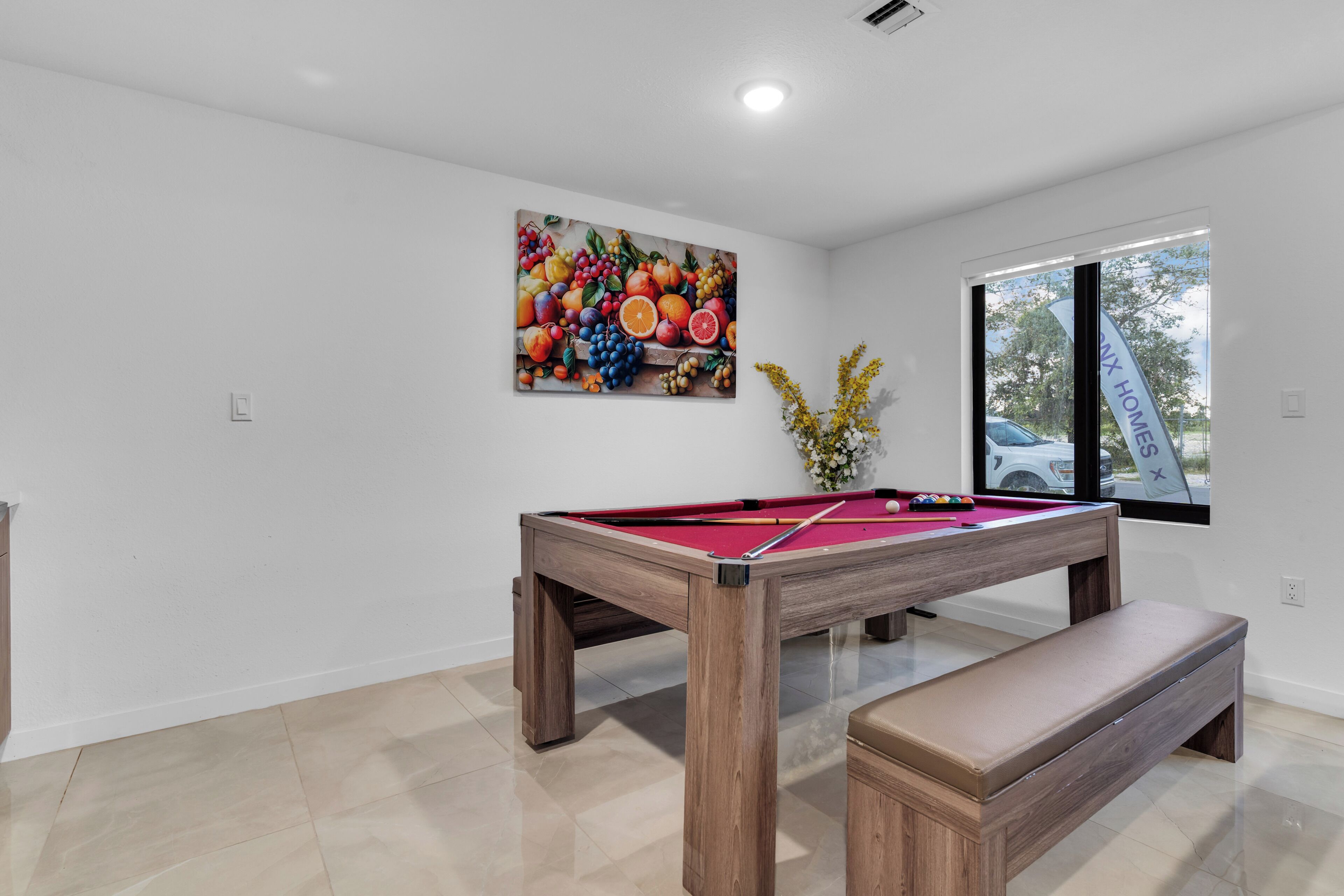 Una mesa de billar con un banco de madera, un cuadro con motivos de frutas en la pared y una ventana con vistas a un coche.
