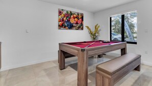 Een biljart met een houten bankje, een schilderij met fruitmotieven aan de muur, en een raam met uitzicht op een auto.